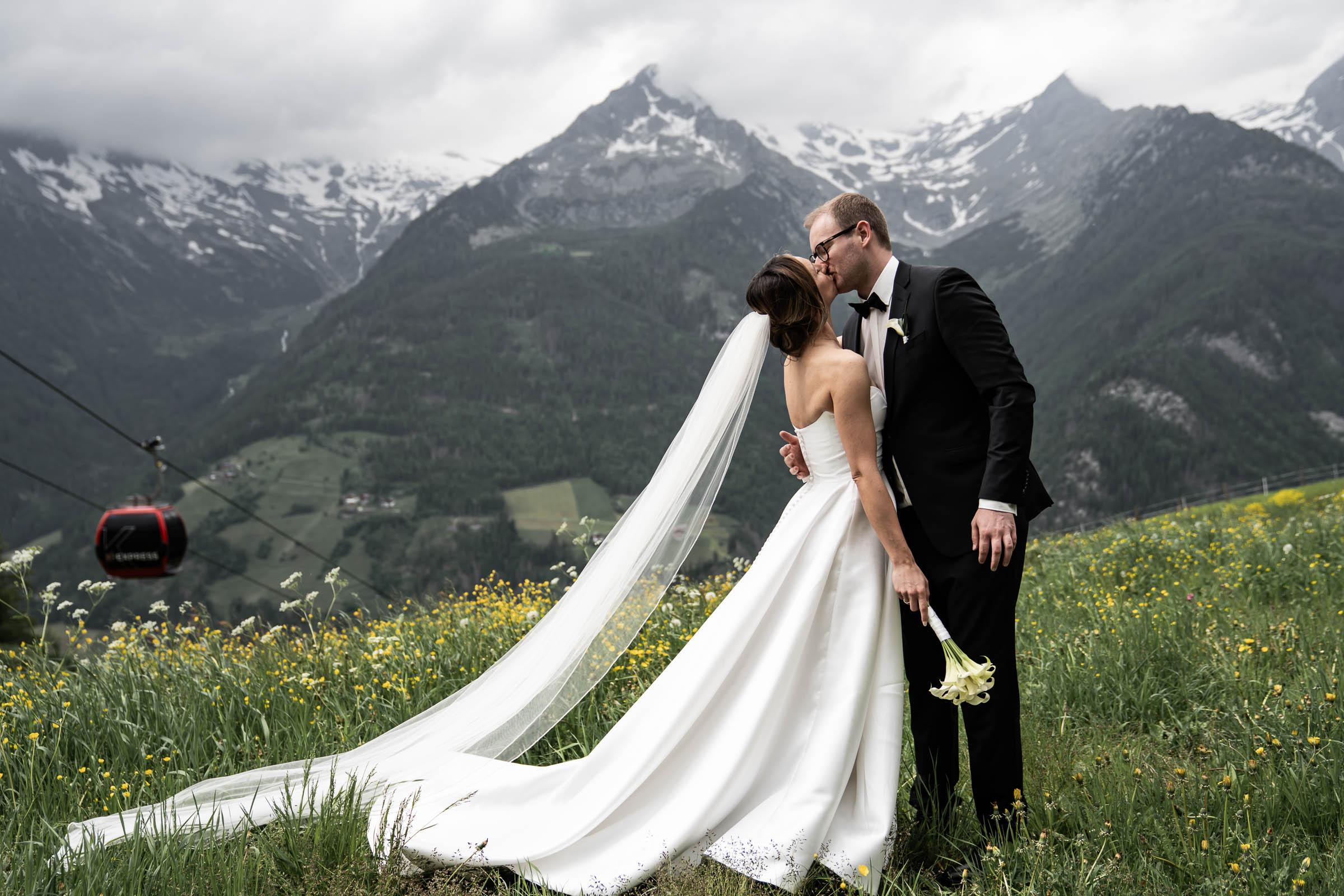 Bride and groom kissing in an alpine meadow, gondola passing behind
