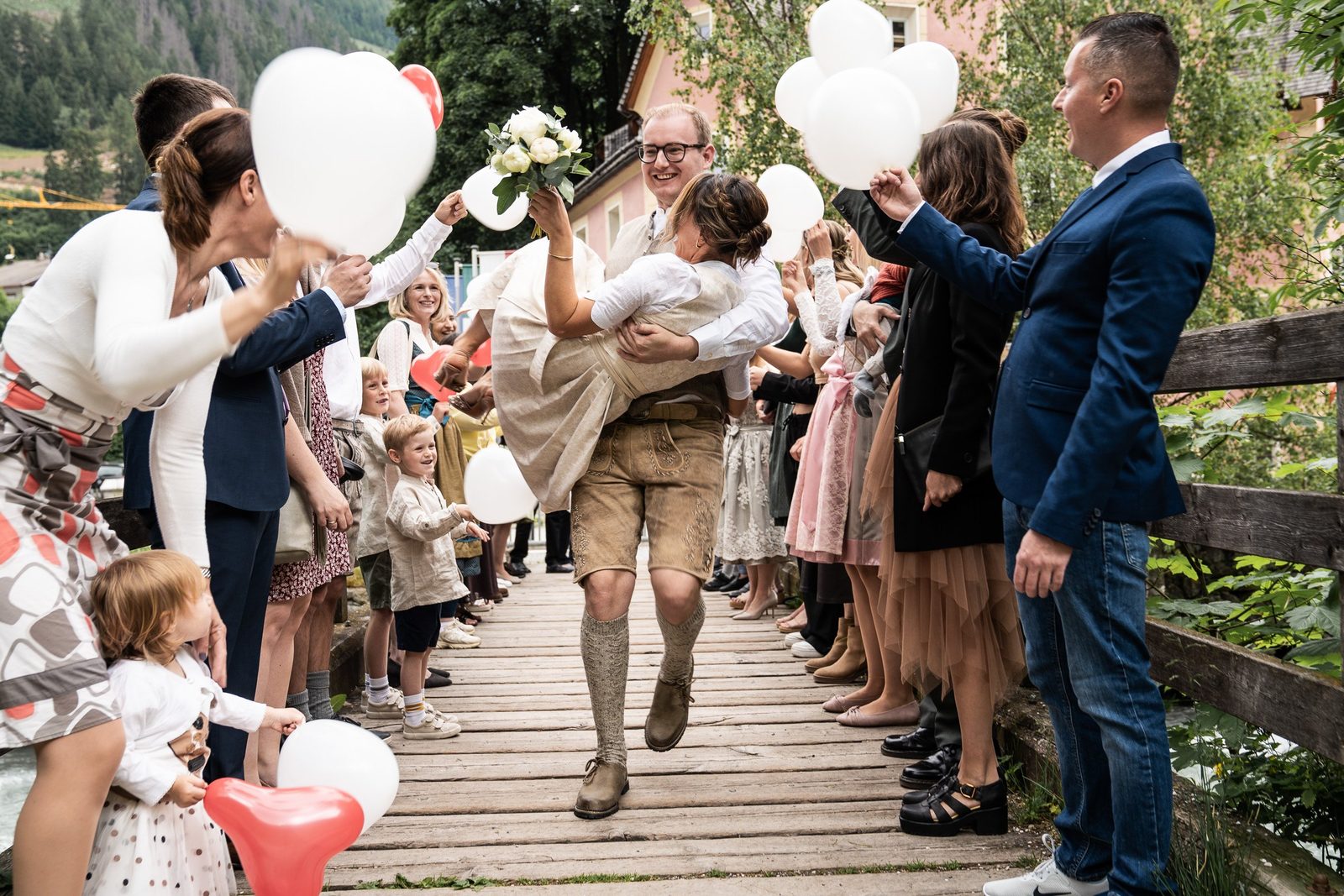 Lo sposo porta in braccio la sposa su una passerella di legno, gli invitati con palloncini bianchi ai lati