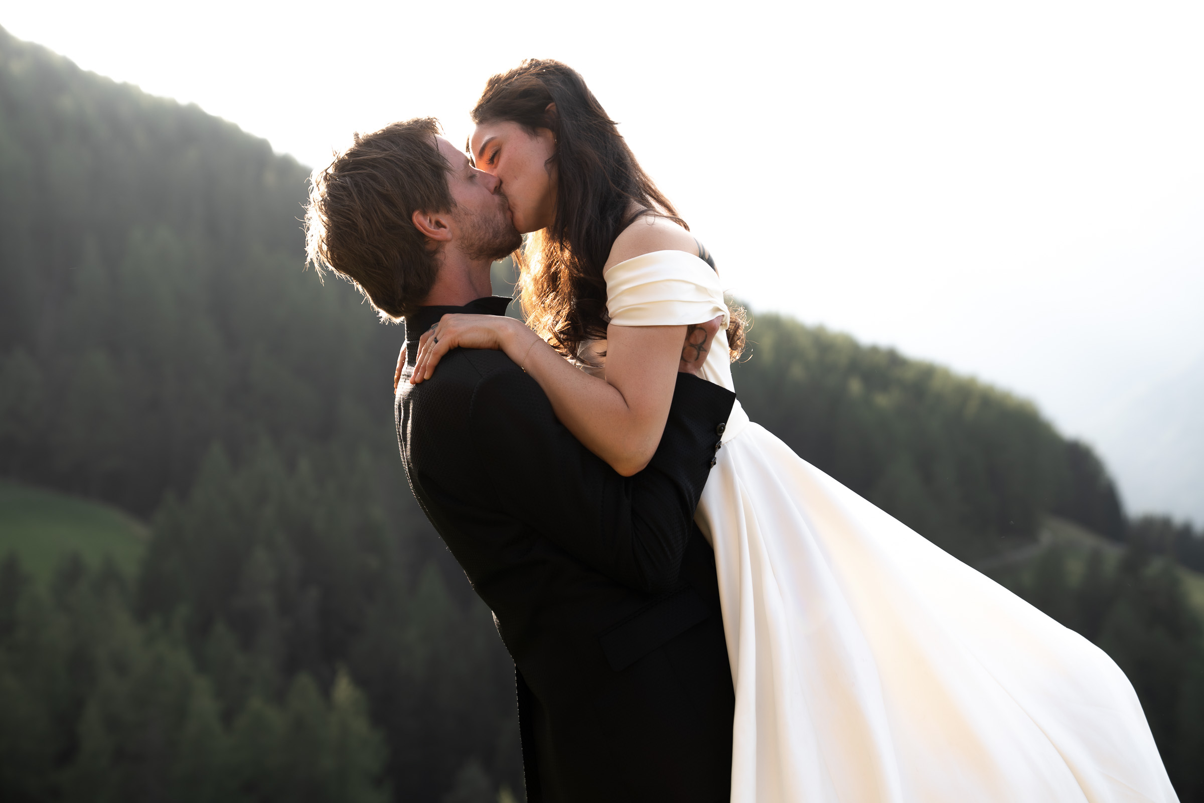 Groom lifting bride in a backlit kiss against forested alpine ridges