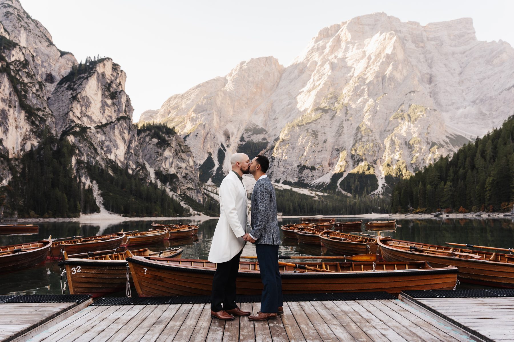 Walker and Joe kiss on the jetty at Lago di Braies, rowing boats and the Seekofel behind
